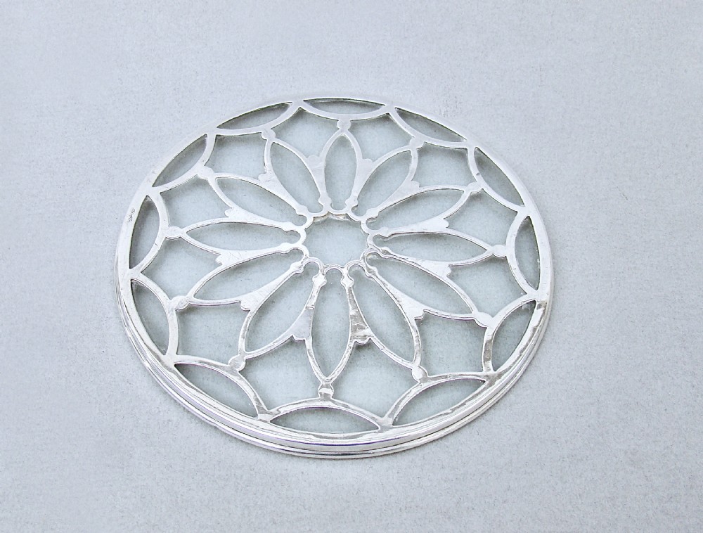 A circular clear glass teapot stand featuring an intricate pierced sterling silver overlay in a petalled geometric pattern.