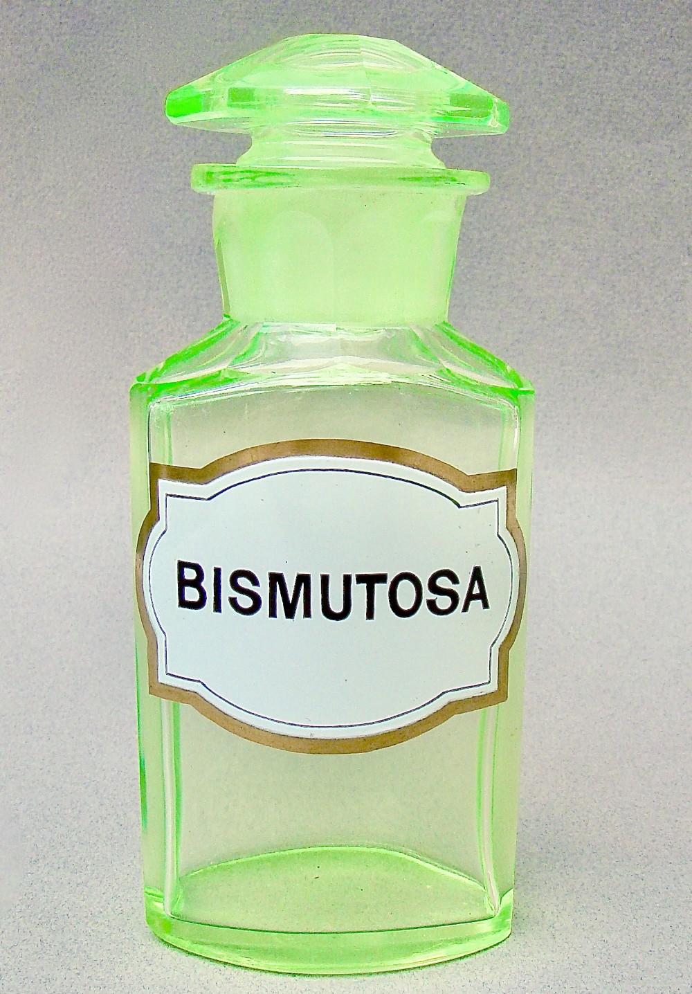 A light-green translucent uranium glass apothecary jar featuring a fitted faceted stopper and a printed label reading Bismutosa.