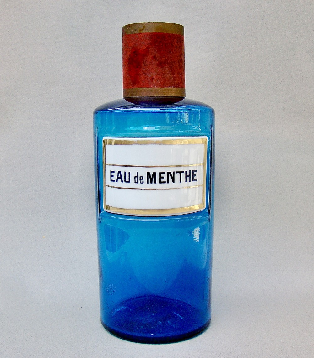 A cylindrical turquoise blue glass apothecary jar featuring a white and gold enamel label reading Eau de Menthe and fitted with a brass and cork stopper.