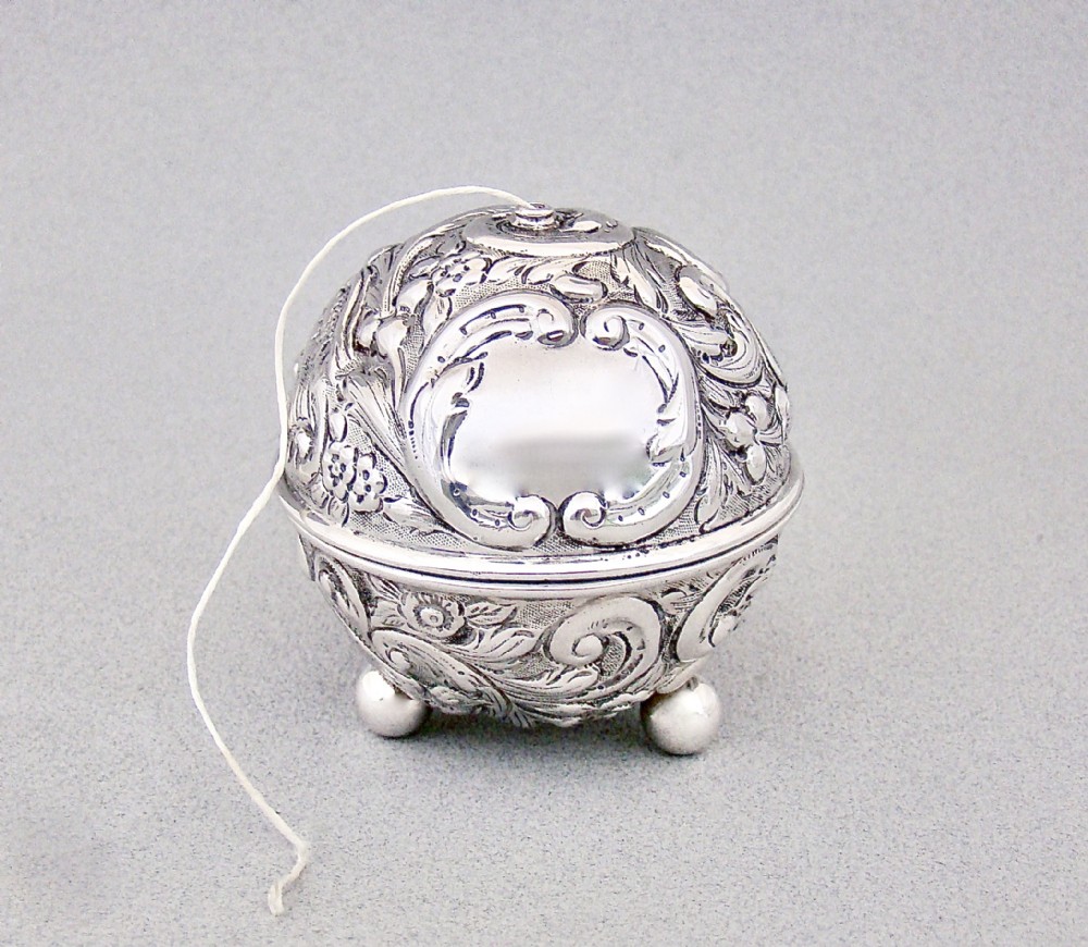 A spherical late Victorian sterling silver string box with chased floral and scroll ornamentation, resting on three ball feet with string extending from the top opening.