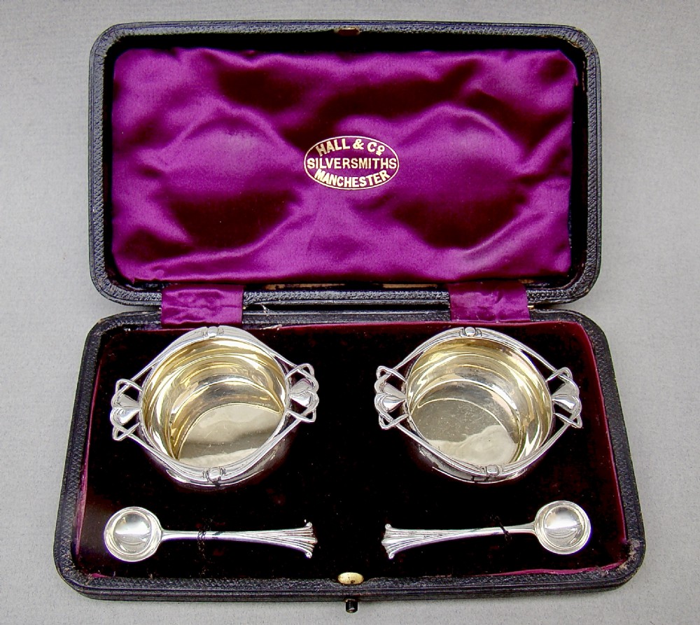 A cased pair of Edwardian sterling silver open salt cellars with stylised handles and matching spoons set in a purple velvet fitted presentation box.