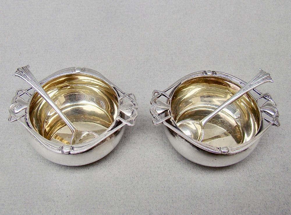 A cased pair of Edwardian sterling silver open salt cellars with stylised handles and matching spoons set in a purple velvet fitted presentation box. View II