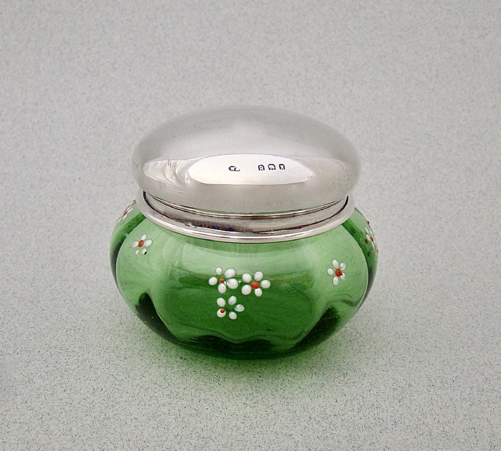 A small green glass jar decorated with white enamel flowers and topped with a hallmarked sterling silver cap.