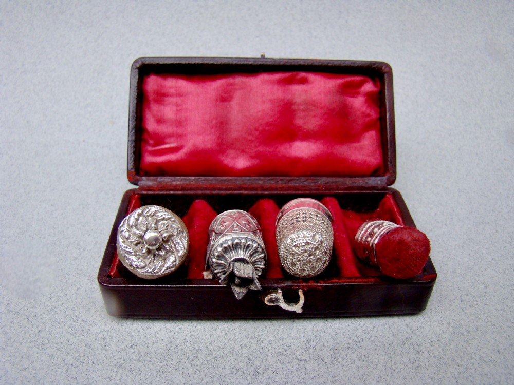 An open dark-cased Victorian sewing set lined with crimson red silk velvet, containing four decorated silver tools including a thimble, needle case, pin cushion, and waxer. View V