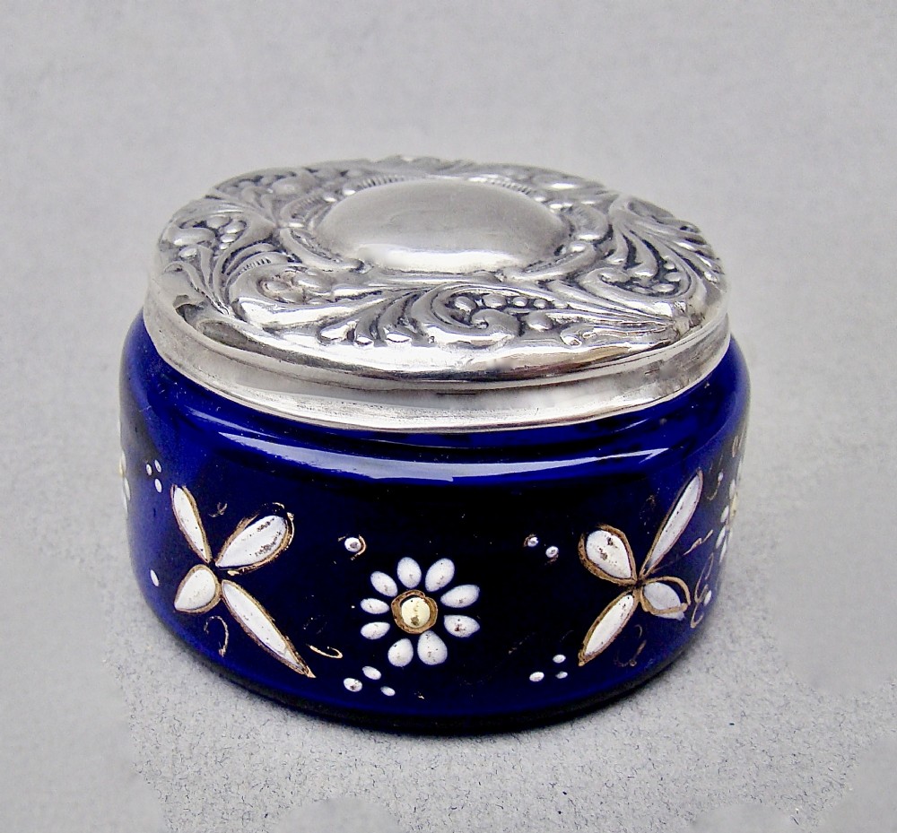 A small cylindrical cobalt blue glass jar decorated with white enamelled floral motifs and topped with an embossed silver lid.