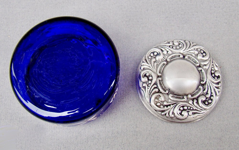 A small cylindrical cobalt blue glass jar decorated with white enamelled floral motifs and topped with an embossed silver lid. View VI