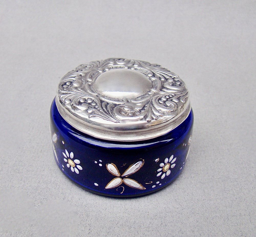 A small cylindrical cobalt blue glass jar decorated with white enamelled floral motifs and topped with an embossed silver lid. View VII
