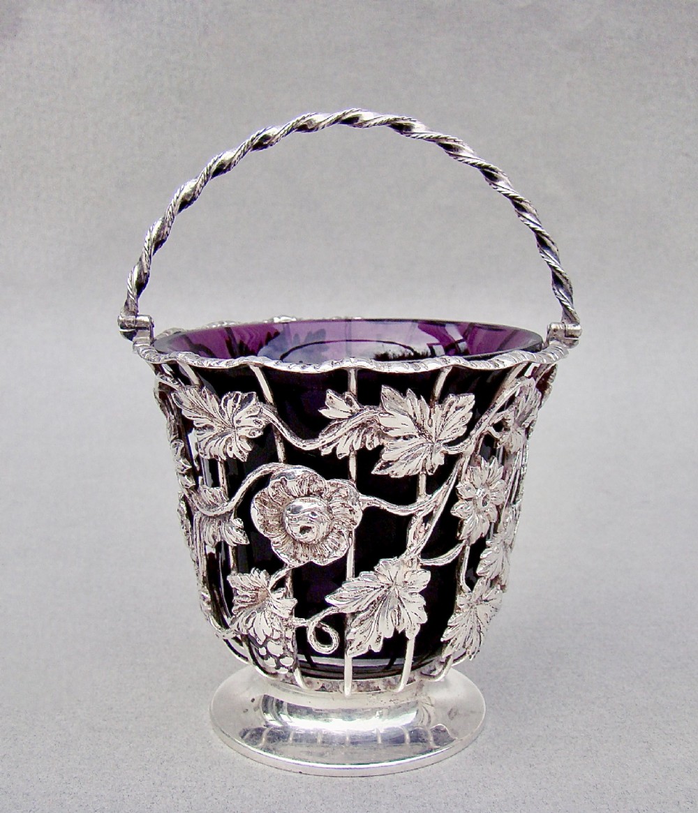 A sterling silver wirework sugar basket featuring a twisted swing handle, cast vine leaf motifs, and a removable purple glass liner.