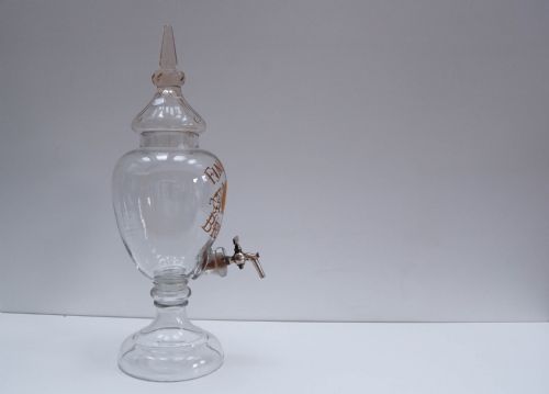 fig 1. A clear glass spirit dispenser featuring gilt typography reading Fine Pale Brandy, a spire finial lid, and a brass spigot at the base.