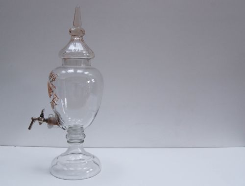 fig 2. A clear glass spirit dispenser featuring gilt typography reading Fine Pale Brandy, a spire finial lid, and a brass spigot at the base.