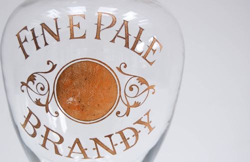 fig 3. A clear glass spirit dispenser featuring gilt typography reading Fine Pale Brandy, a spire finial lid, and a brass spigot at the base.