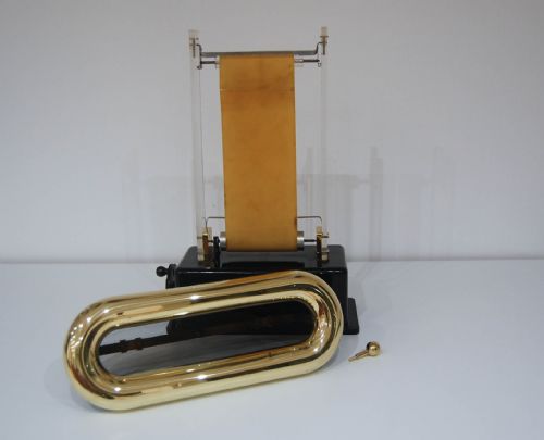 fig 2. A hand-cranked Van de Graaff electrostatic generator featuring a black metal base, transparent acrylic supports, an upright rubber belt, and a wire mesh upper dome trimmed in brass.