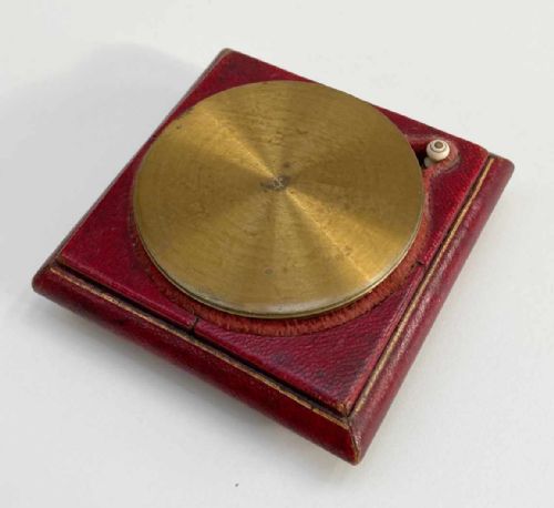 fig 1. A square red leather desk compass with a circular silvered dial, brass bezel, and a separate brass protective cap.
