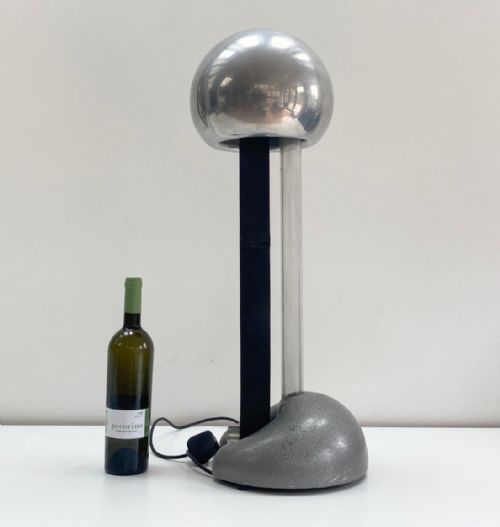 fig 2. A mid-twentieth-century Van de Graaff generator featuring a polished silver electrostatic sphere supported on vertical columns above a grey textured base, accompanied by a discharge wand with a wooden handle.