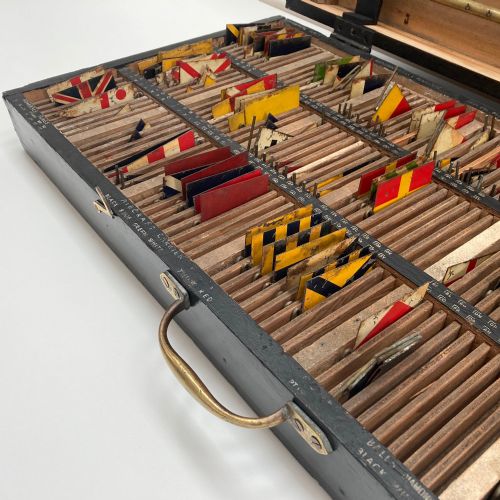 fig 2. An open early twentieth-century black wooden box featuring internal compartments with miniature signal flags and upright brass masts displaying colourful painted flags.