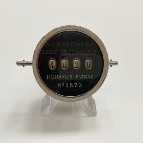 fig 1. A late Victorian pocket revolution counter made of nickel metal with a round glass face, resting inside its open original dark brown leather case lined with bright purple velvet.