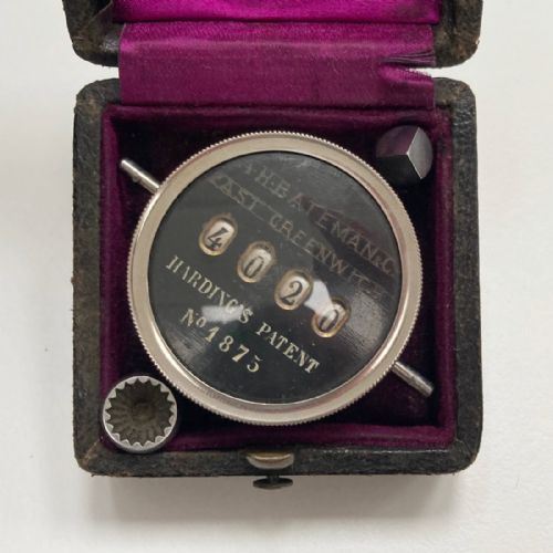 fig 8. A late Victorian pocket revolution counter made of nickel metal with a round glass face, resting inside its open original dark brown leather case lined with bright purple velvet.