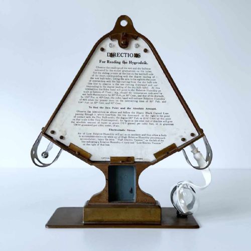 fig 6. A triangular brass Hygrodeik scientific hygrometer with glass tube thermometers on both sides and a central calculating chart with a pointer.