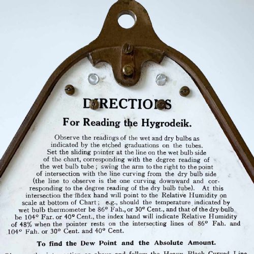 fig 7. A triangular brass Hygrodeik scientific hygrometer with glass tube thermometers on both sides and a central calculating chart with a pointer.