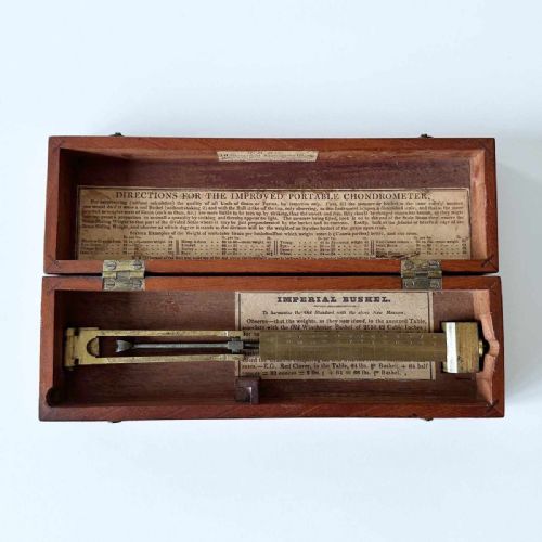 fig 2. An antique brass portable chondrometer scale featuring a small bucket suspended from a graduated beam, resting within a fitted mahogany case with printed paper labels.