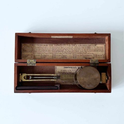 fig 3. An antique brass portable chondrometer scale featuring a small bucket suspended from a graduated beam, resting within a fitted mahogany case with printed paper labels.