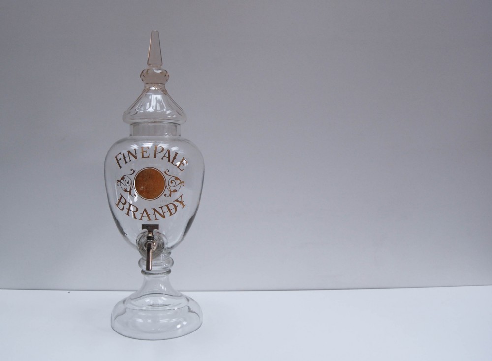 A clear glass spirit dispenser featuring gilt typography reading Fine Pale Brandy, a spire finial lid, and a brass spigot at the base.