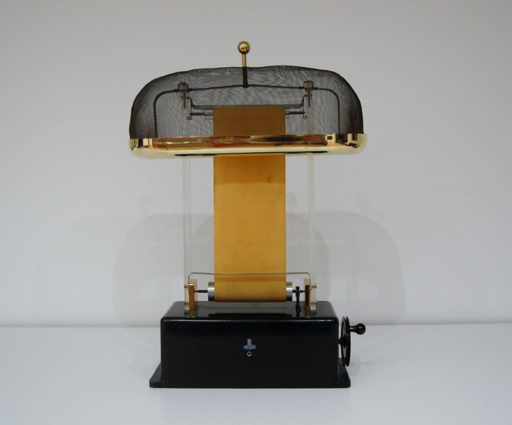 A hand-cranked Van de Graaff electrostatic generator featuring a black metal base, transparent acrylic supports, an upright rubber belt, and a wire mesh upper dome trimmed in brass.