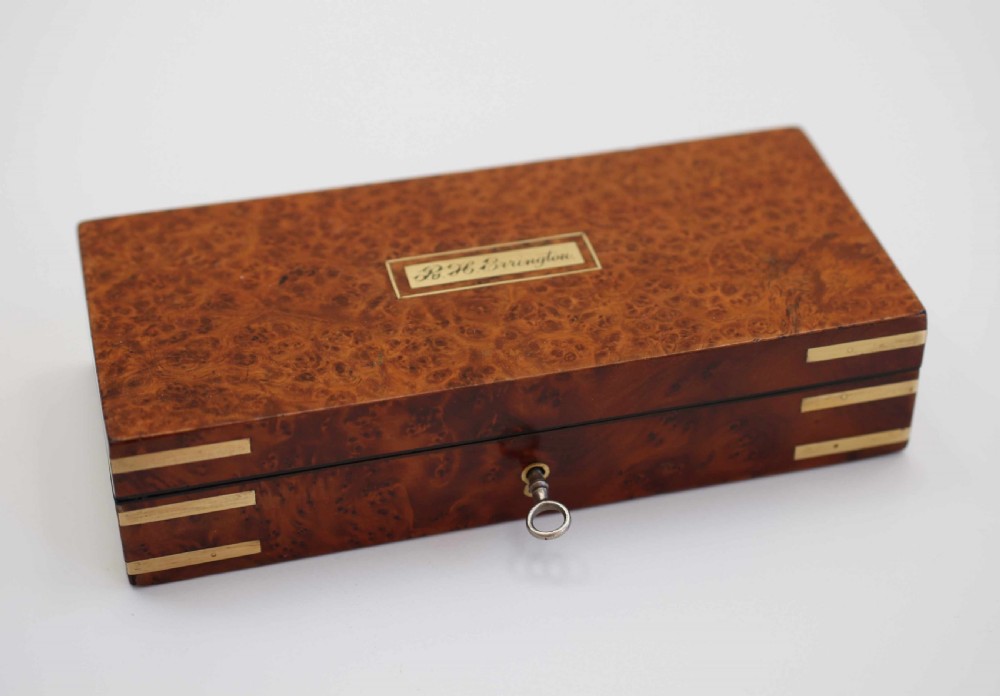 A rectangular burr walnut box featuring brass corner straps, a brass nameplate cartouche on the lid, and a key inserted into the front keyhole.