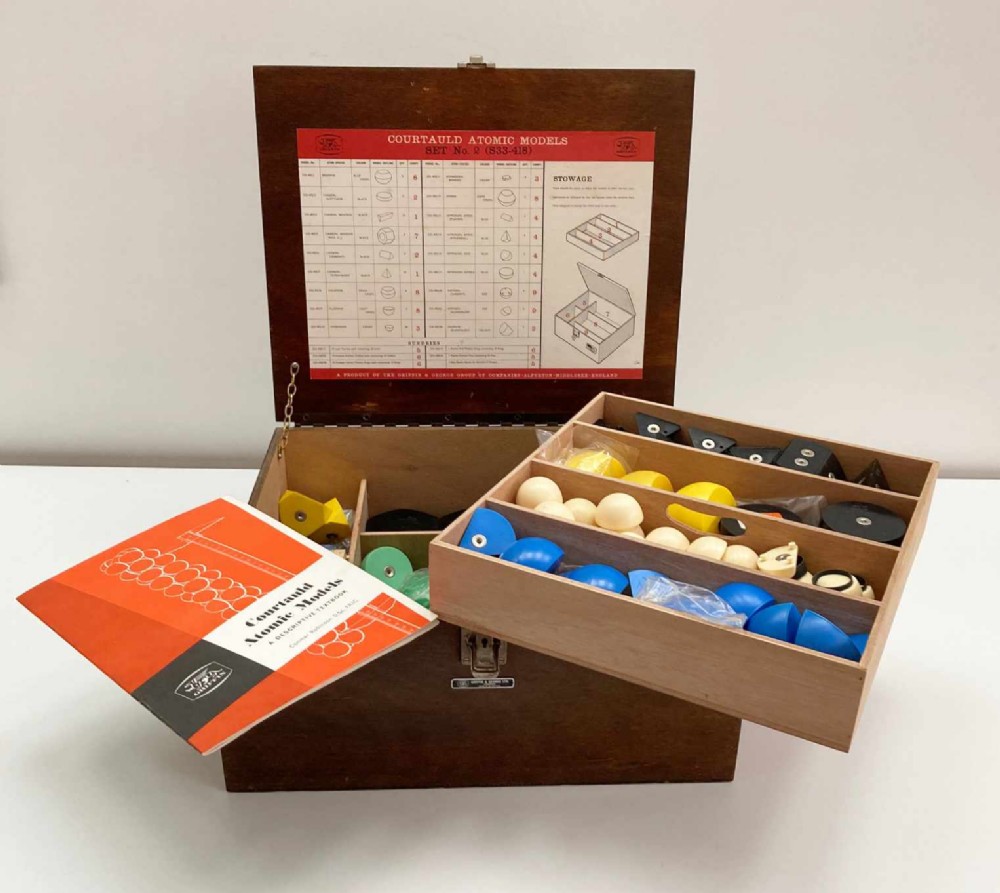A vintage wooden case filled with colourful plastic atomic model components, accompanied by an instructional booklet and a printed chart inside the lid.