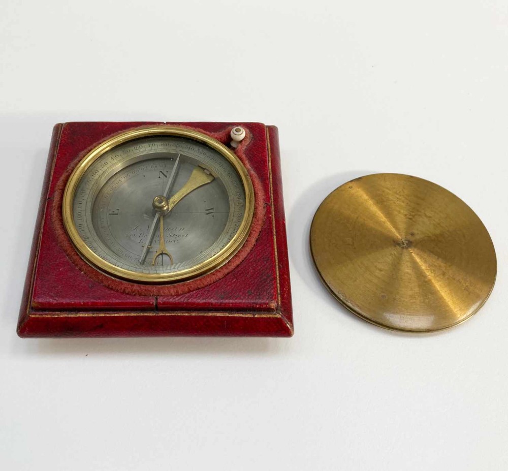 A square red leather desk compass with a circular silvered dial, brass bezel, and a separate brass protective cap.