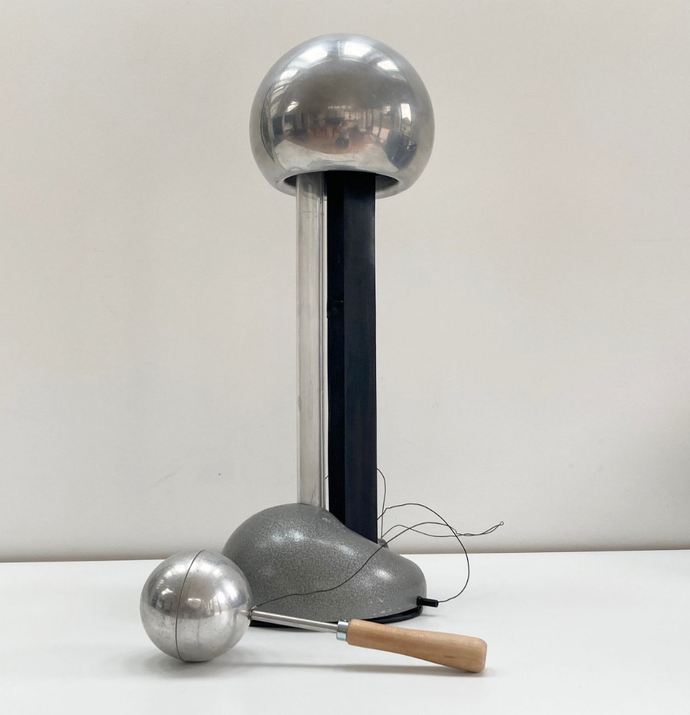 A mid-twentieth-century Van de Graaff generator featuring a polished silver electrostatic sphere supported on vertical columns above a grey textured base, accompanied by a discharge wand with a wooden handle.