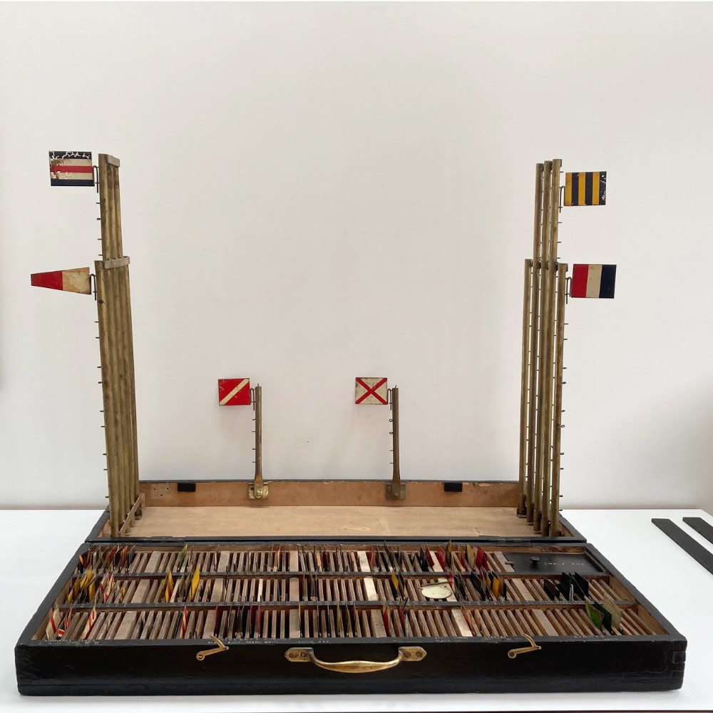 An open early twentieth-century black wooden box featuring internal compartments with miniature signal flags and upright brass masts displaying colourful painted flags.