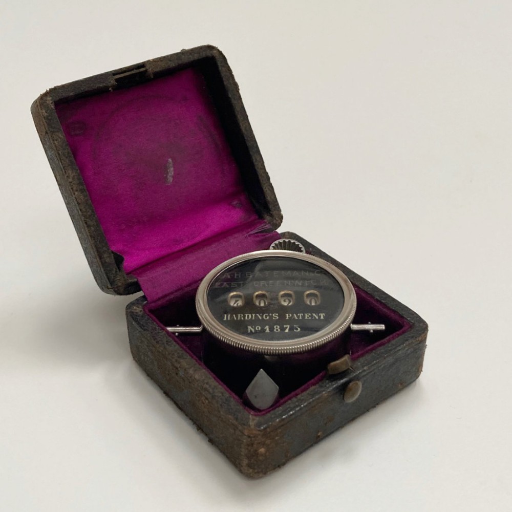 A late Victorian pocket revolution counter made of nickel metal with a round glass face, resting inside its open original dark brown leather case lined with bright purple velvet.