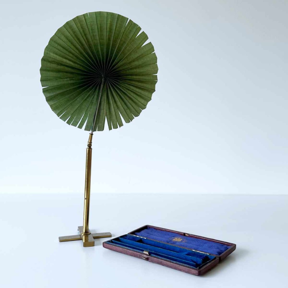 An antique Victorian brass microscopy candle shade with a pleated green fan screen standing beside its original velvet-lined presentation case.