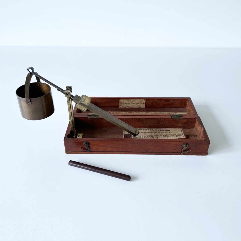 An antique brass portable chondrometer scale featuring a small bucket suspended from a graduated beam, resting within a fitted mahogany case with printed paper labels.