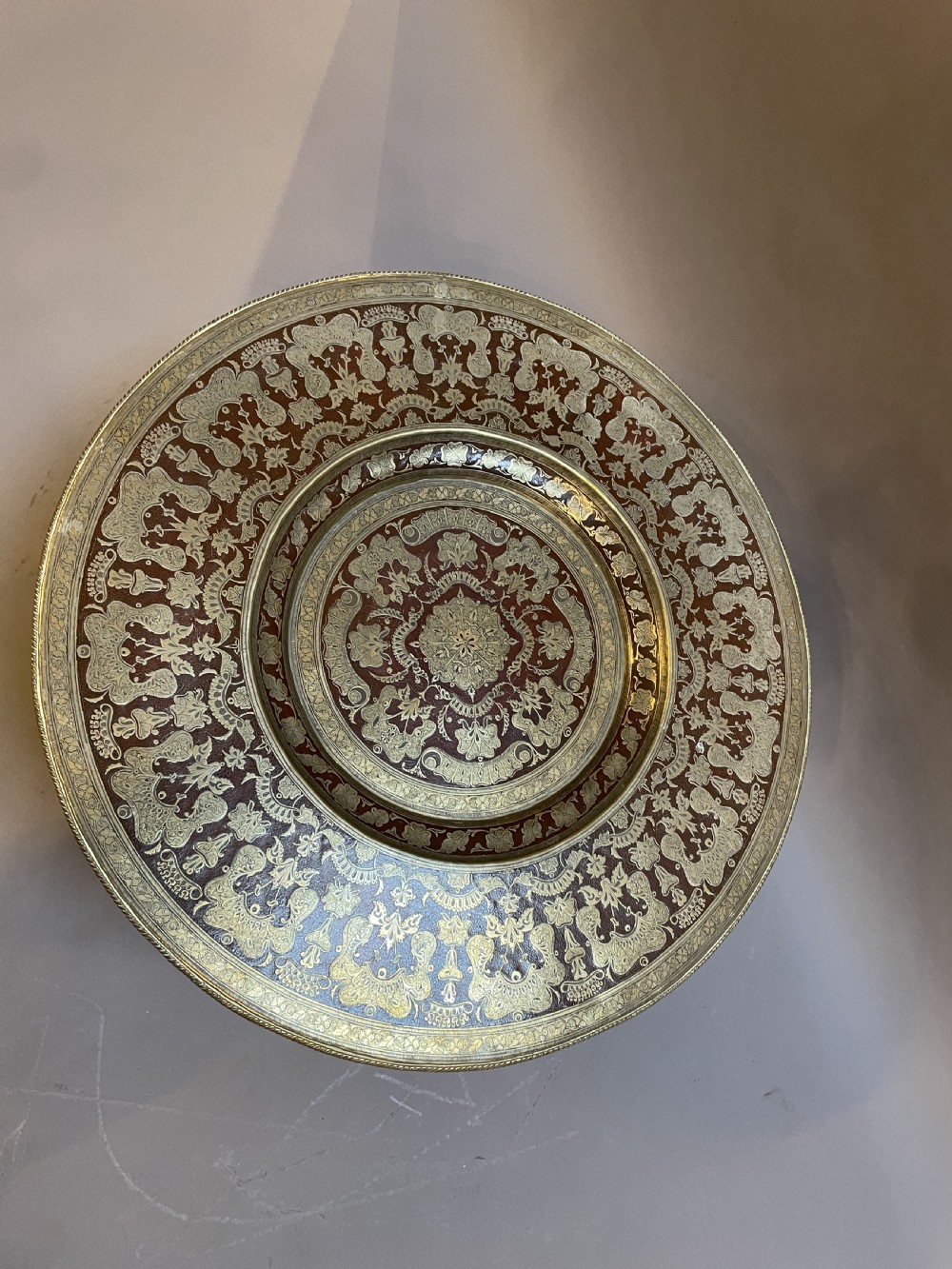 A low circular occasional table featuring an intricately etched multi-metal tray top resting on a dark carved wooden base. View IX