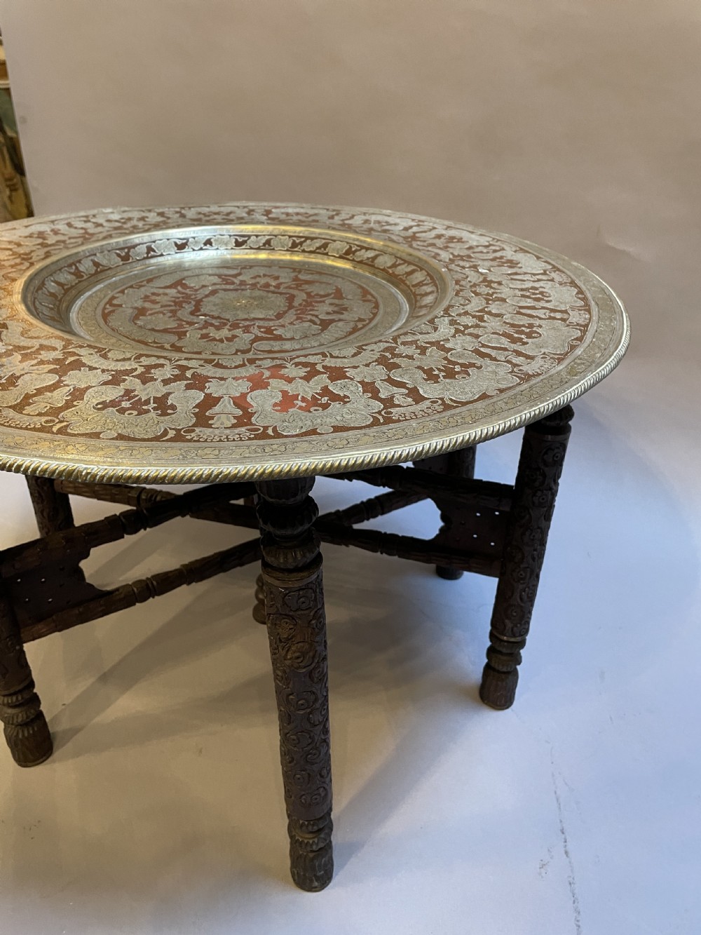 A low circular occasional table featuring an intricately etched multi-metal tray top resting on a dark carved wooden base. View X