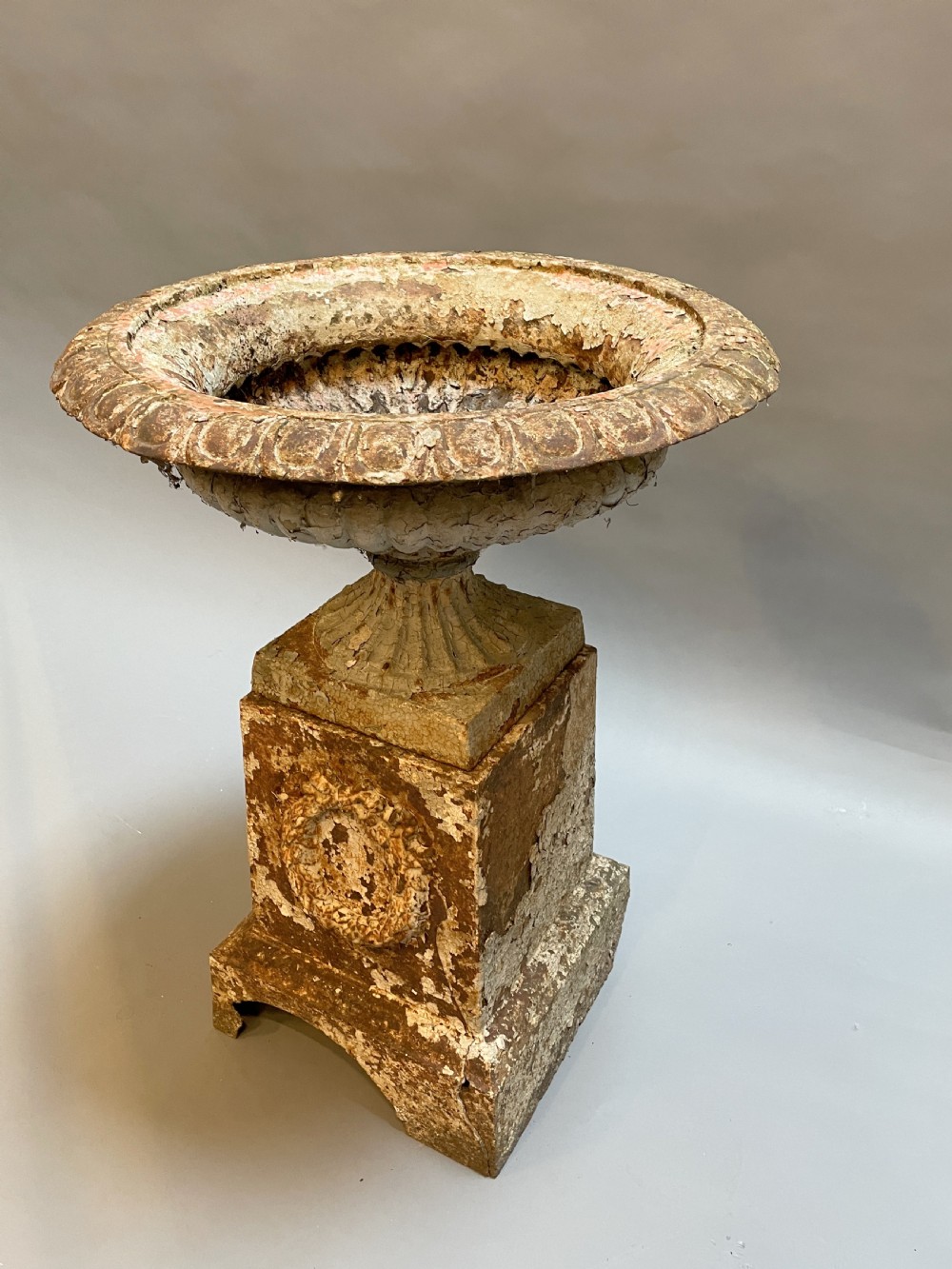 A Victorian cast iron garden urn with a fluted rim and laurel wreath medallion base, displaying a weathered white paint finish over oxidised iron.