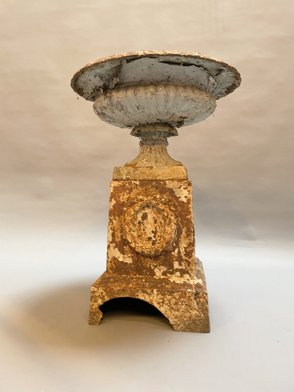 A Victorian cast iron garden urn with a fluted rim and laurel wreath medallion base, displaying a weathered white paint finish over oxidised iron. View IV
