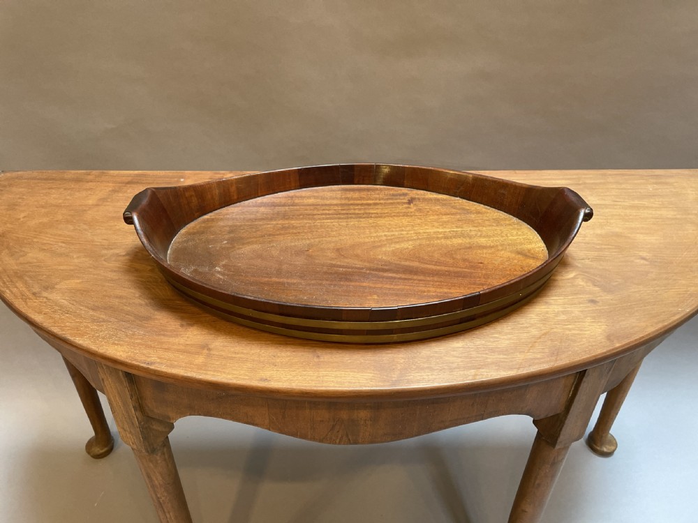 A Georgian Revival oval mahogany serving tray featuring a raised rim, integrated handles, and a brass band outer trim.