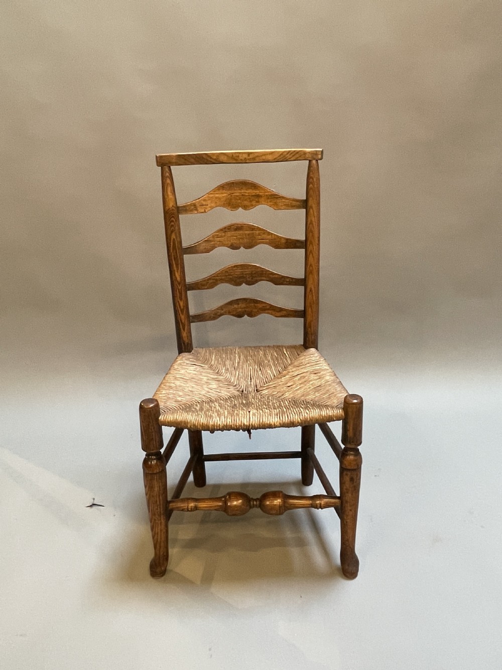 A wooden country side chair featuring a ladderback with four wavy horizontal slats, turned front legs, and a woven rush seat.