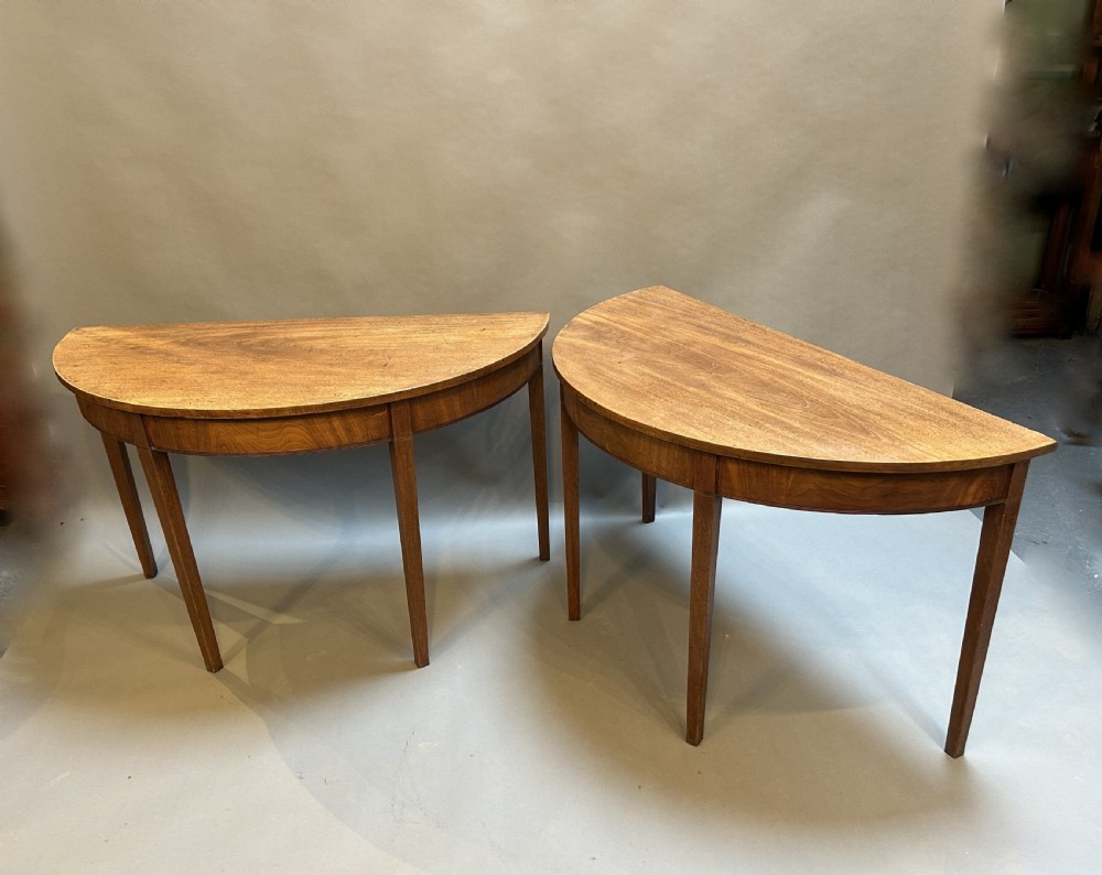 A pair of George III mahogany semi-circular demilune console tables supported by square tapered legs.