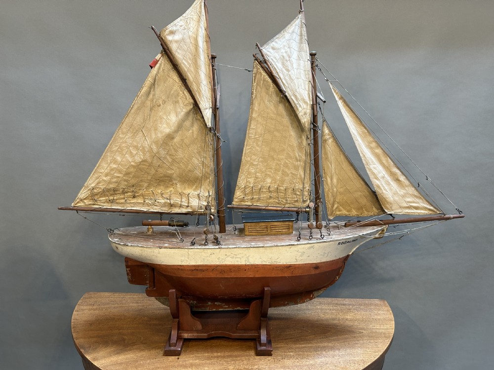 A large vintage wooden pond yacht with a two-masted schooner rig, aged sails, and a white and brown painted hull mounted on a dark wooden stand.