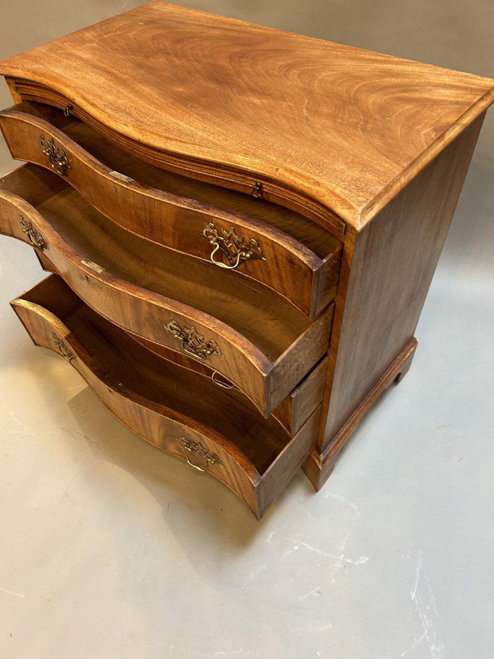 A 19th-century mahogany chest of drawers featuring a serpentine front, four graduated drawers with ornamental brass handles, and bracket feet. View VII