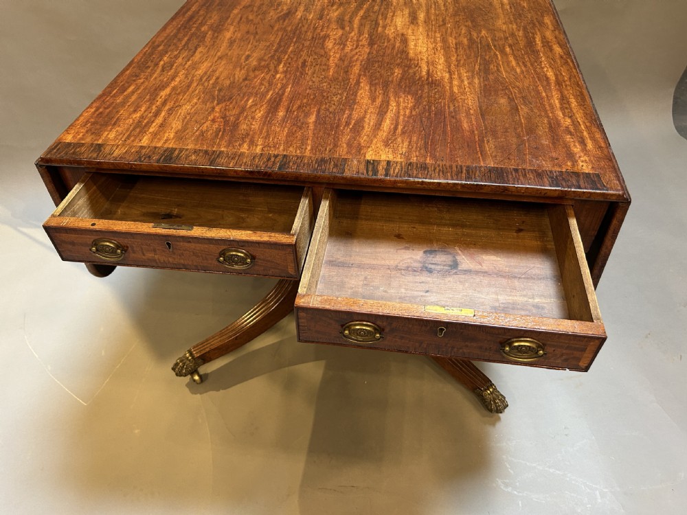 A George IV mahogany sofa table featuring drop leaves, frieze drawers with brass handles, and a reeded quadruped pedestal base with brass castors. View VI