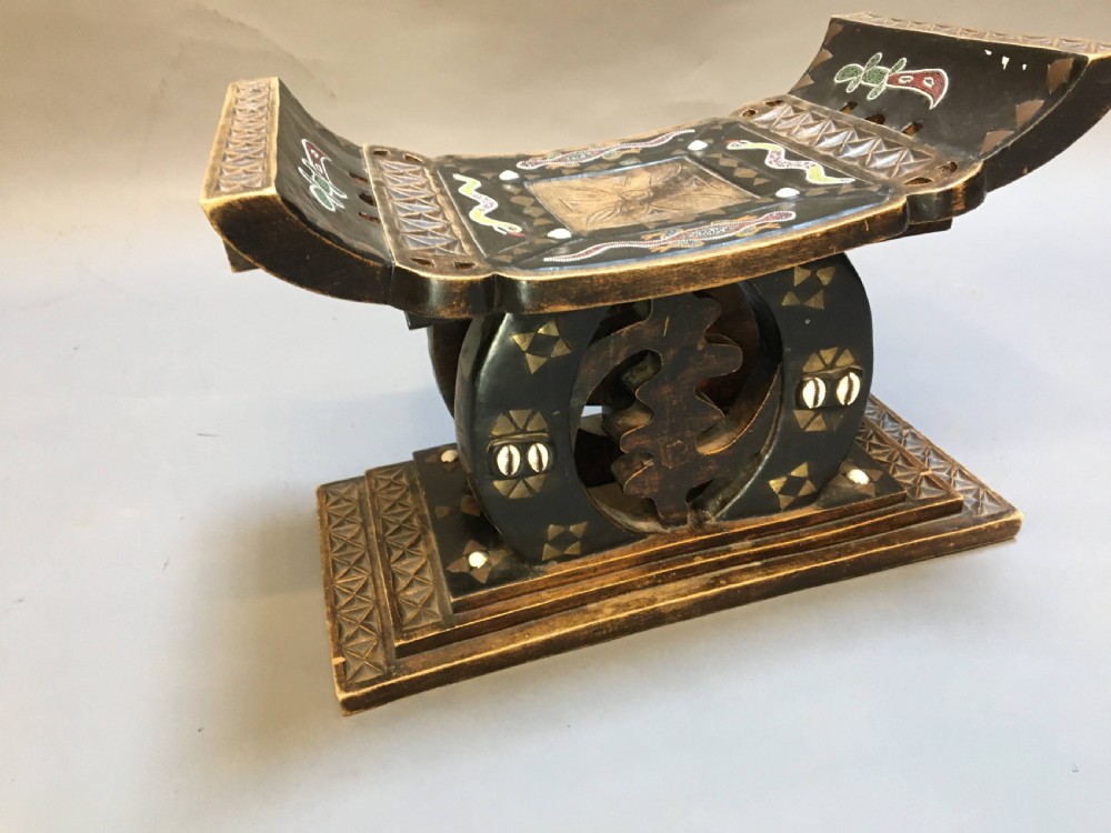 A hand-carved dark timber Ashanti stool featuring a curved seat with inlaid motifs, a central openwork column, and a rectangular carved base. View V
