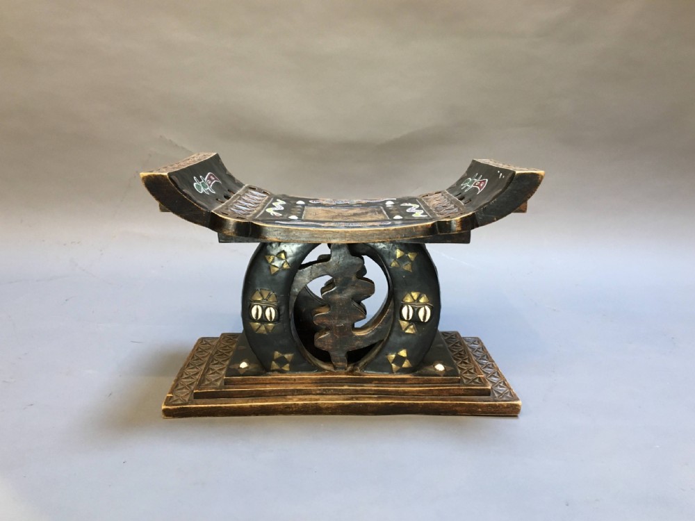A hand-carved dark timber Ashanti stool featuring a curved seat with inlaid motifs, a central openwork column, and a rectangular carved base. View VI