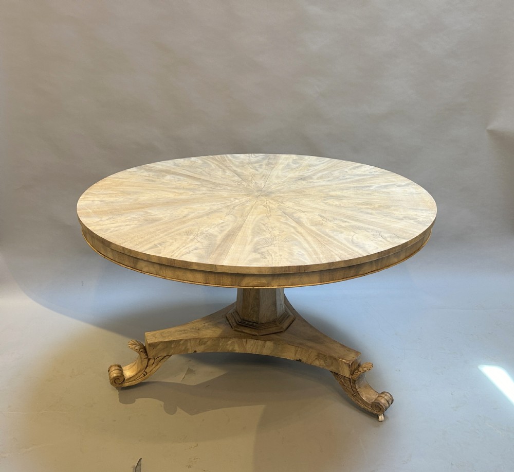 A 19th-century circular dining table featuring a radially veneered top, a faceted central pedestal column, and a carved triform base with scroll feet.