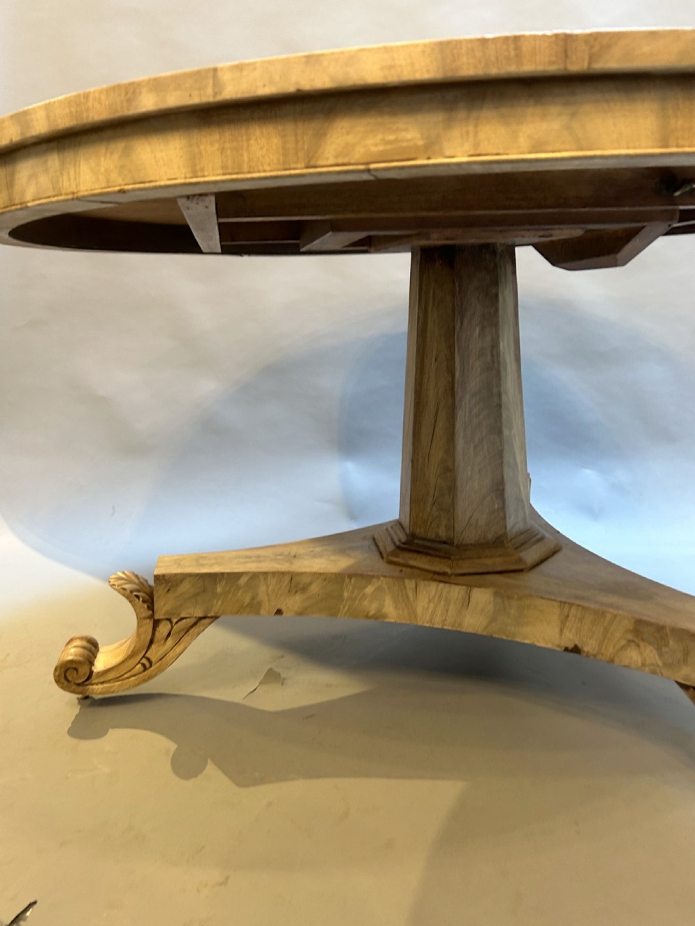 A 19th-century circular dining table featuring a radially veneered top, a faceted central pedestal column, and a carved triform base with scroll feet. View VI