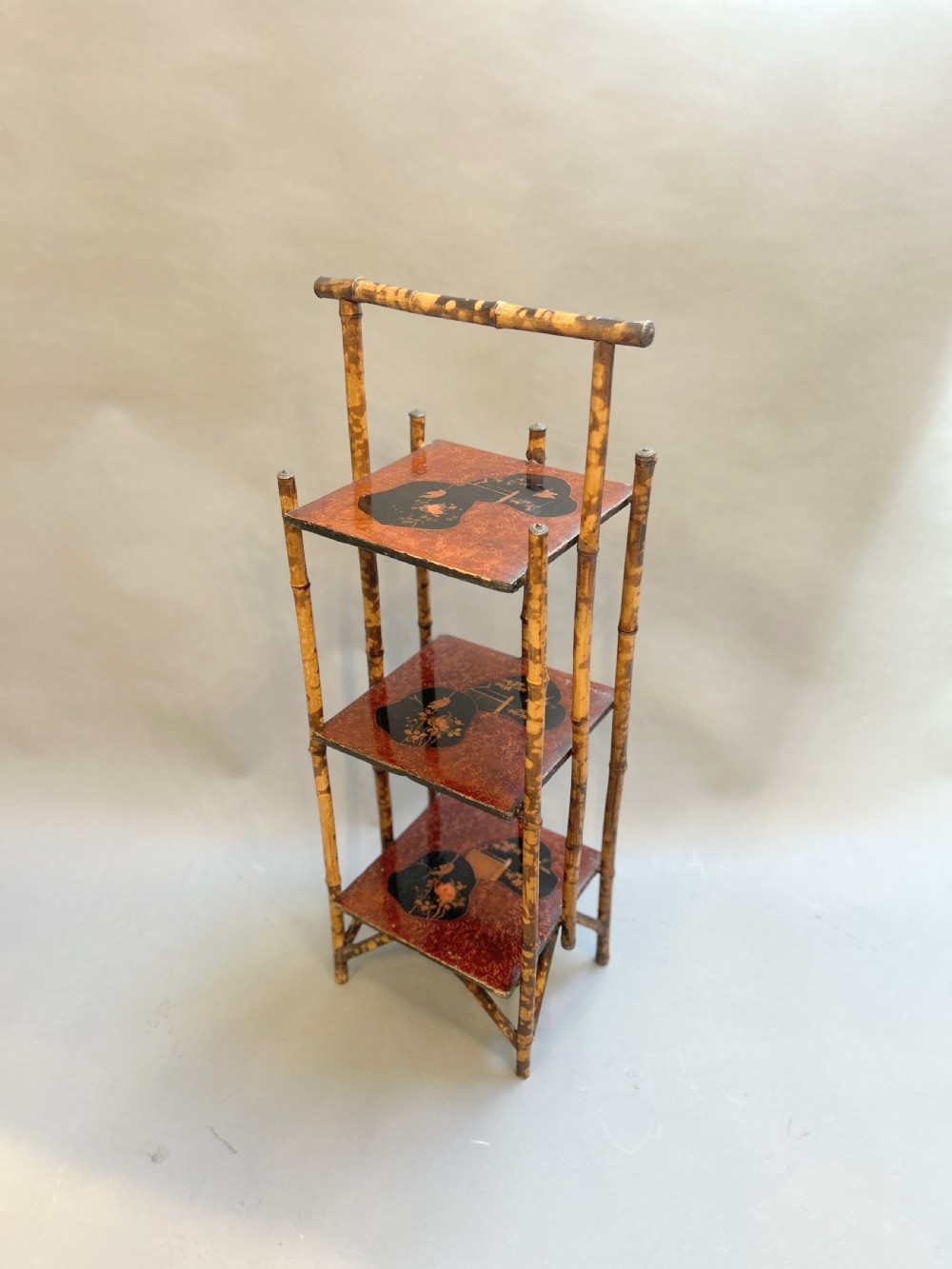 A three-tiered late Victorian bamboo side table featuring a mottled finish, square lacquered shelves decorated with fan motifs, and an overarching top handle.
